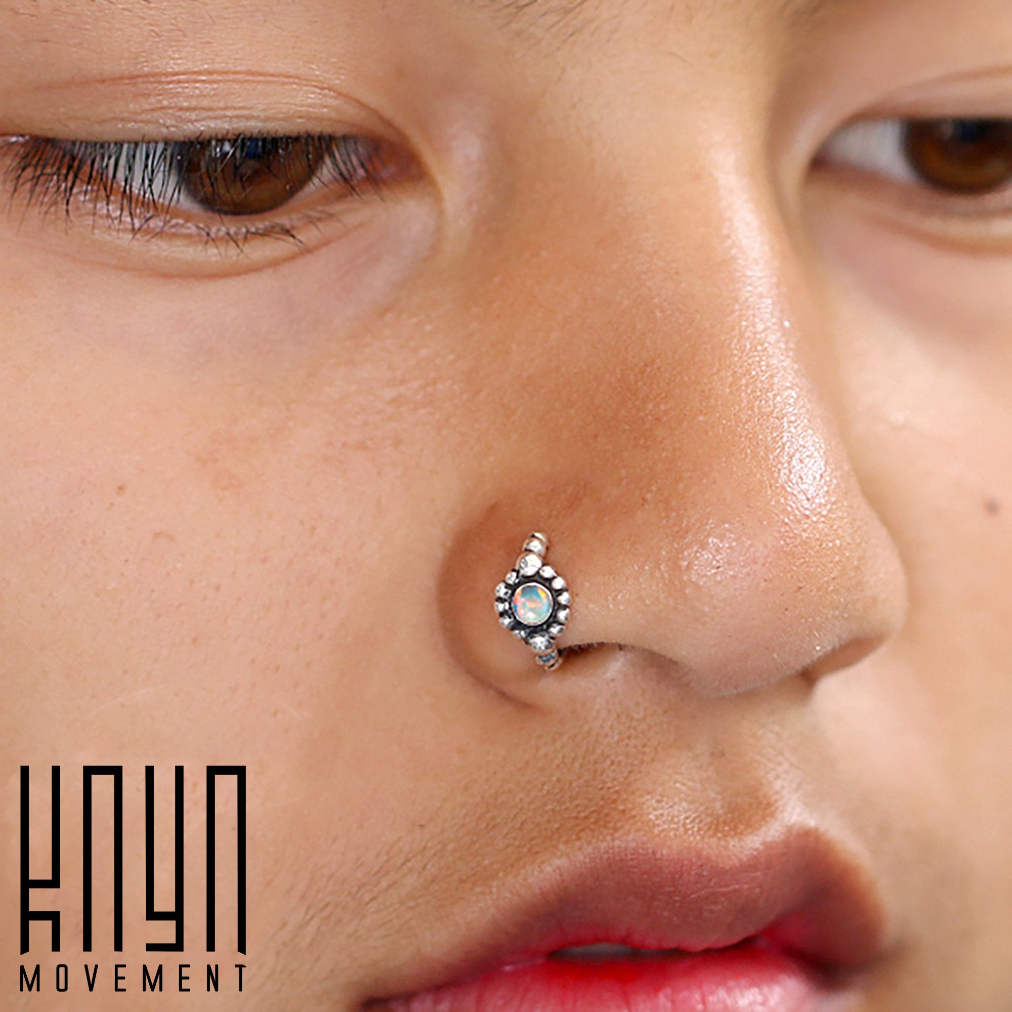Close-up image of a person with a nose piercing featuring Kaya Movement's SHA Beaded Flower Nose Ring in Silver & White Opal, crafted in 20 gauge. The skin around the piercing is smooth and blemish-free. The text "Kaya Movement" appears in the lower left corner.